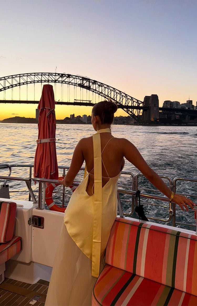 Rear view of a yellow satin halter neck maxi dress with an open back, thin criss-cross straps, and a wide vertical scarf deta