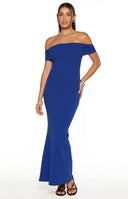 A rear three-quarter view of our model wearing a form-fitting floor-length in a vibrant blue maxi dress features a wide off-the-shoulder neckline with delicate ruching on the sleeves. This maxi has a straight column silhouette that accentuates her figure.