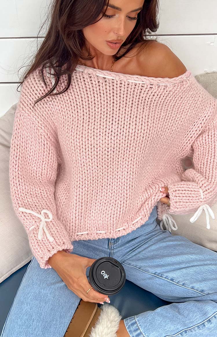 Off-shoulder baby pink chunky knit sweater with white ribbon lacing on the neckline and hem, and white ribbon bows on the cuffs