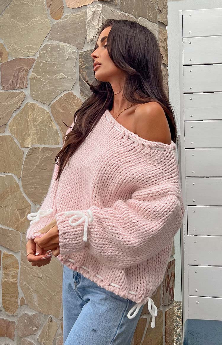 Side view of a baby pink chunky knit sweater with an off-shoulder neckline and white tie details on the cuffs and hem
