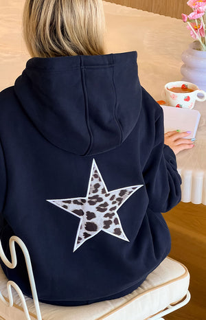 The back view of a navy blue hoodie featuring a large white star appliqué with a cow print pattern on the back.