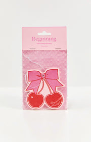 Two red cherry air fresheners with a pink bow and white string, in pink packaging with a BB logo pattern