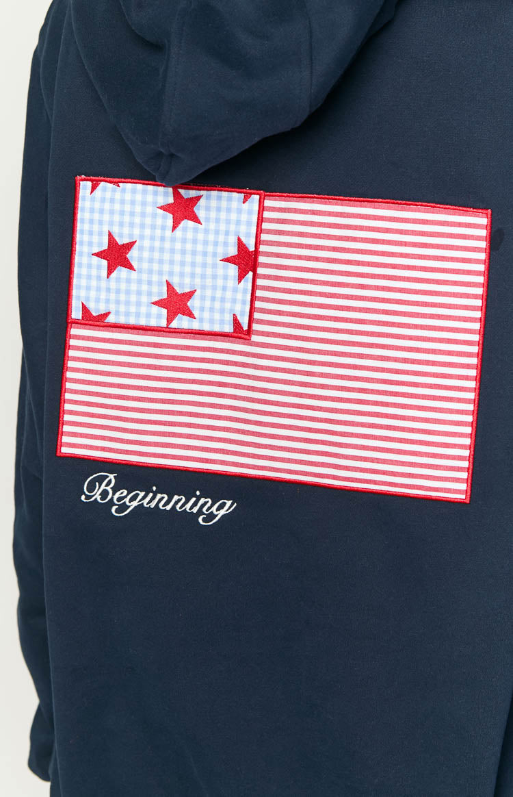 Close-up of the back of a navy hoodie with an embroidered US flag patch, red stripes, blue gingham stars, and white 'Beginning' script