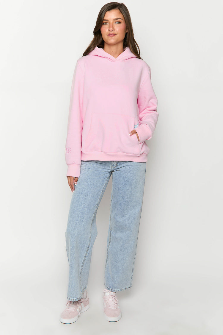 Full-length view of a light pink oversized hoodie with a hood, long sleeves, kangaroo pocket, and blue embroidery on the pocket