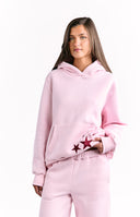 A side view of an oversized light pink hoodie featuring a large dark red star applique. The hoodie has a large front pocket and is part of a matching set.