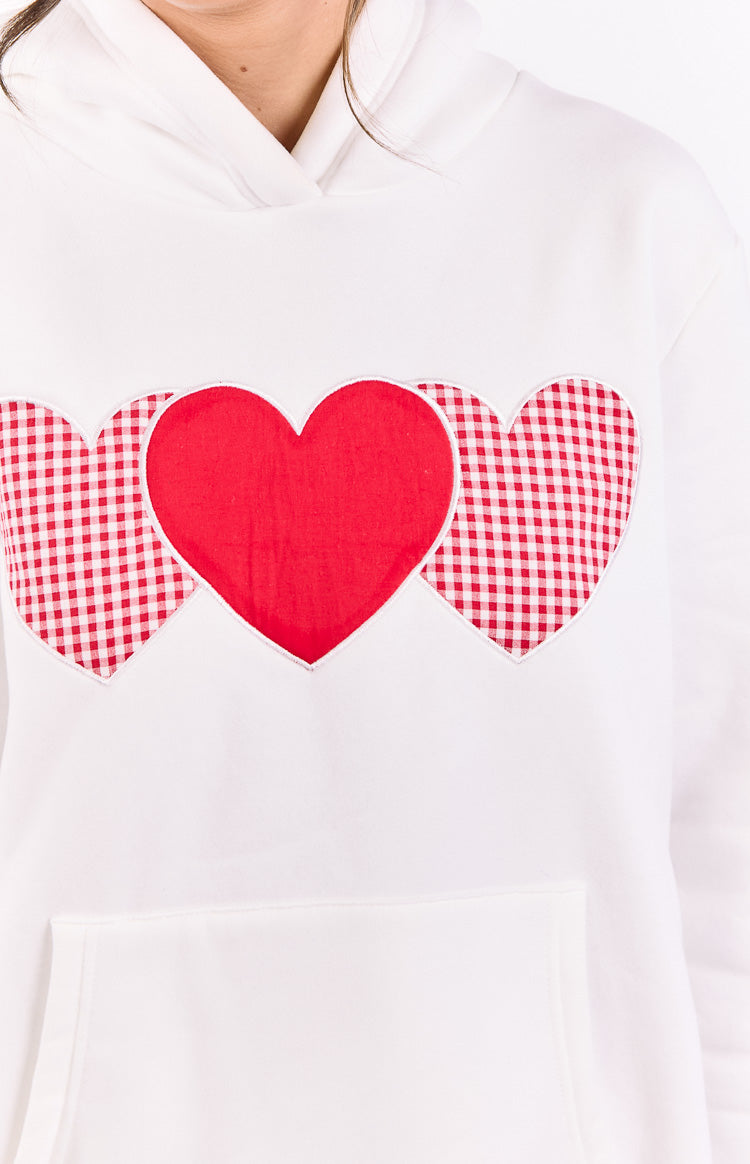 Front detail of a white hoodie with three heart appliques, a central red heart and two red gingham hearts, above a kangaroo pocket