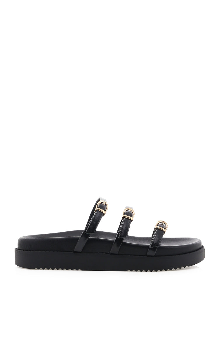 Side view of black slides with three patent leather straps, gold buckles, and a thick ridged platform sole