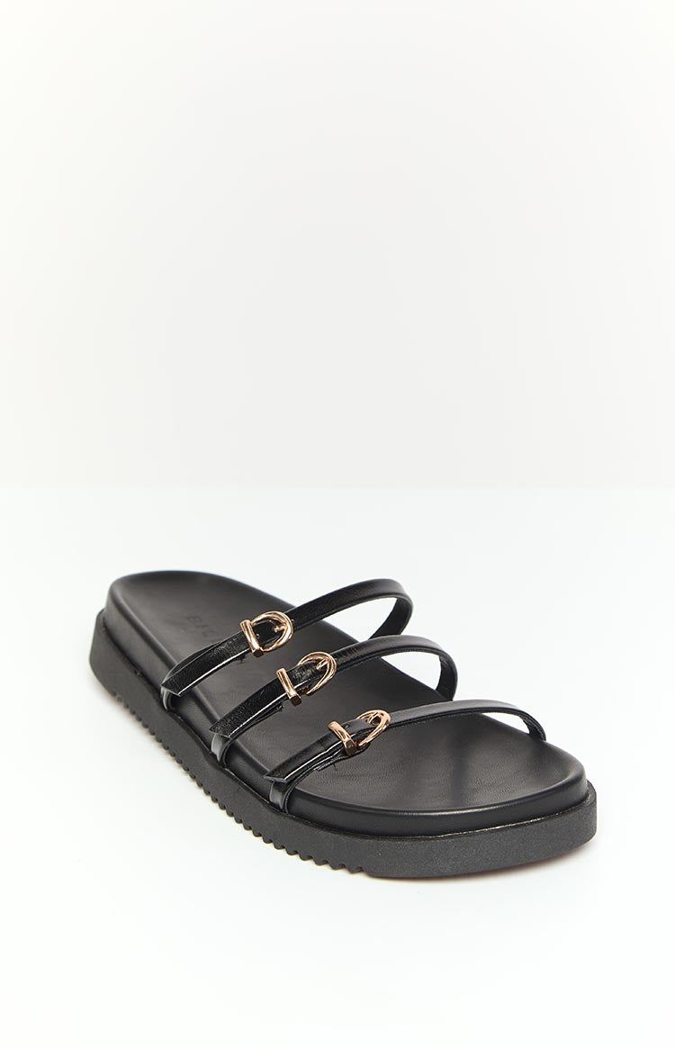 Side view of a black slide showing three patent leather straps with gold buckle details and a thick ridged sole