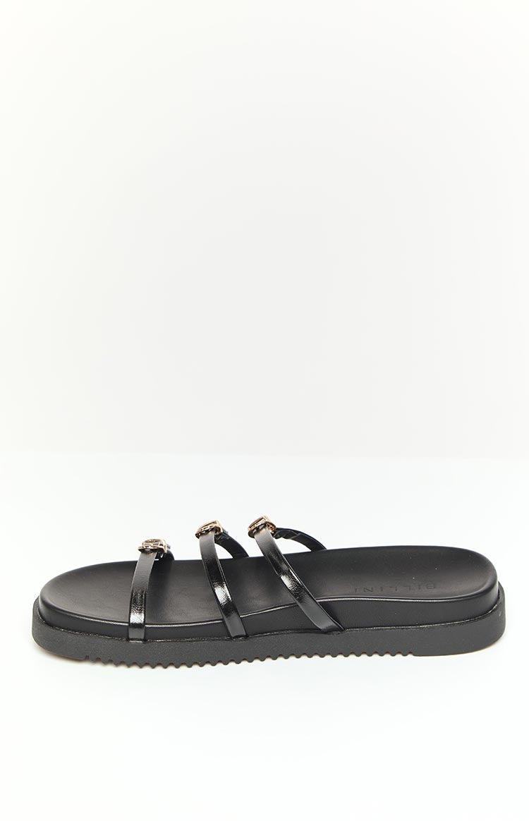 Side view of a black slide with three thin straps and gold buckles on a thick ridged sole