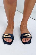 Top-down view of black slides with a square toe and a wide strap featuring a gold abstract buckle