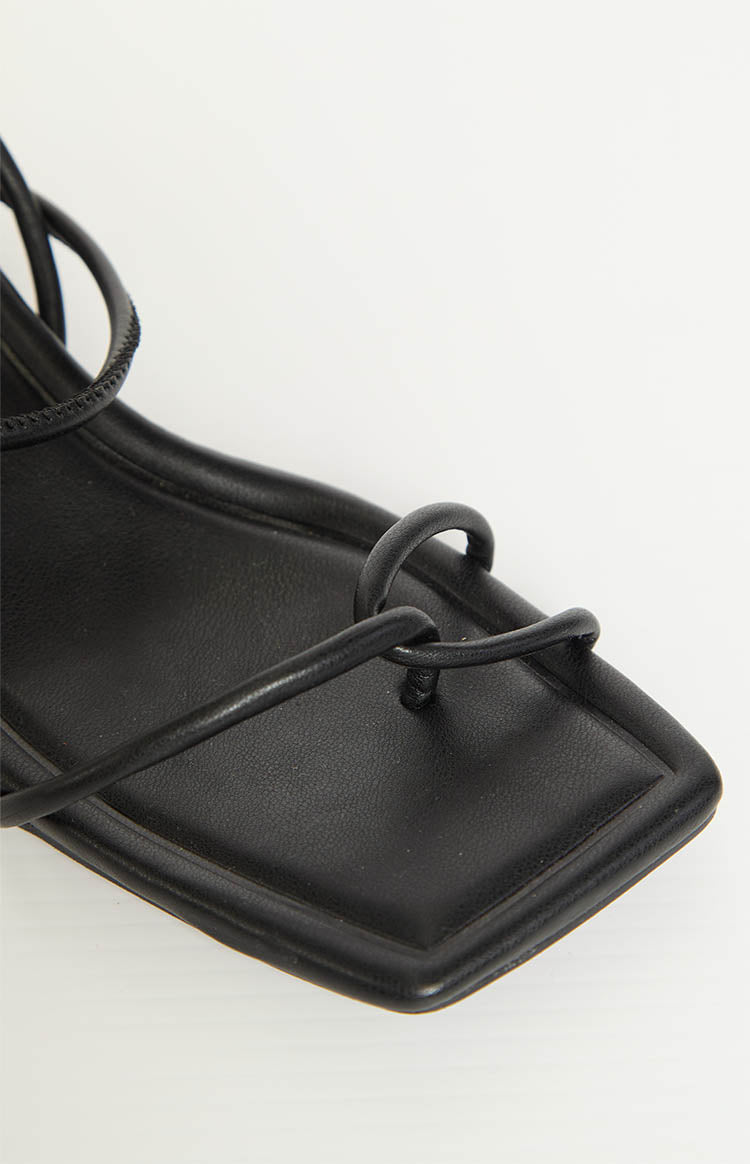 Close-up showing the square toe, thin thong strap, and delicate ankle straps of black heels