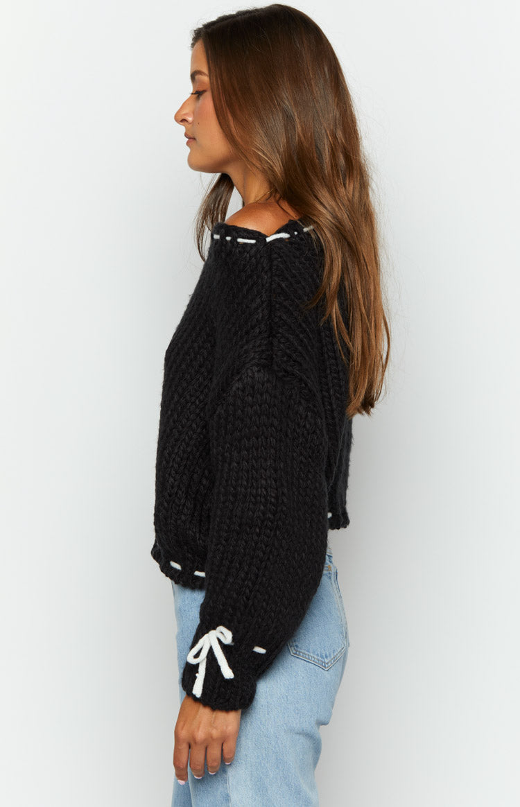 Side view of a black chunky knit off-shoulder sweater with white stitching along the neckline and a white bow on the cuff