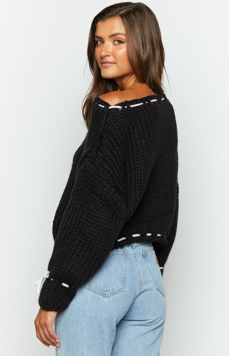 Side view of a black chunky knit sweater with an off-shoulder neckline, white stitching detail, and white cuff ties
