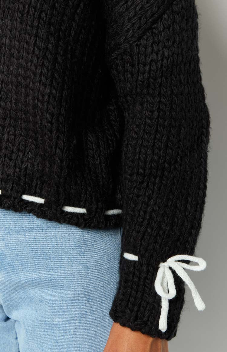 Close-up of the black chunky knit sweater showing white tie detail on the cuff and white stitching along the hem