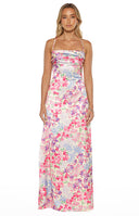 A full-length studio shot of our model wearing a floor-length satin-style maxi dress. This garment features an all-over vibrant floral print with pink purple and green blossoms on a light-colored background. It has thin spaghetti straps a straight-across neckline and a horizontally ruched bodice.