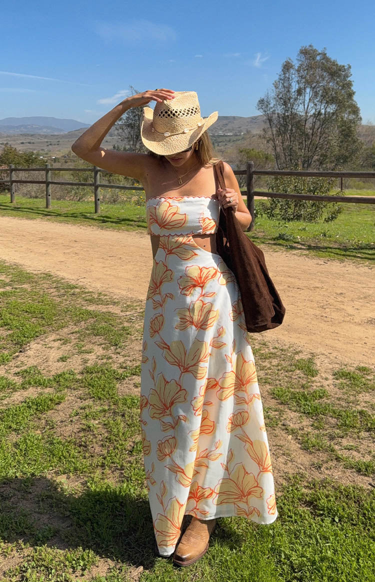 Full length view of a white strapless maxi dress with orange and yellow floral print, waist cutouts, and a scalloped bodice edge