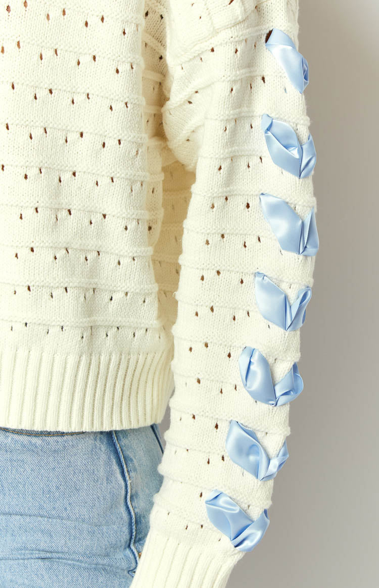 Close-up of the cream knit sweater sleeve with blue satin ribbons tied along the arm, eyelet pattern, and ribbed cuff