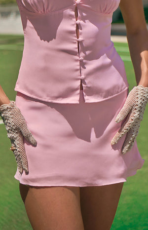 A lifestyle shot from the waist to the mid-thigh of our model wearing a short light pink mini skirt. This mini skirt has a smooth simple design and is paired with a matching pink button-up top.