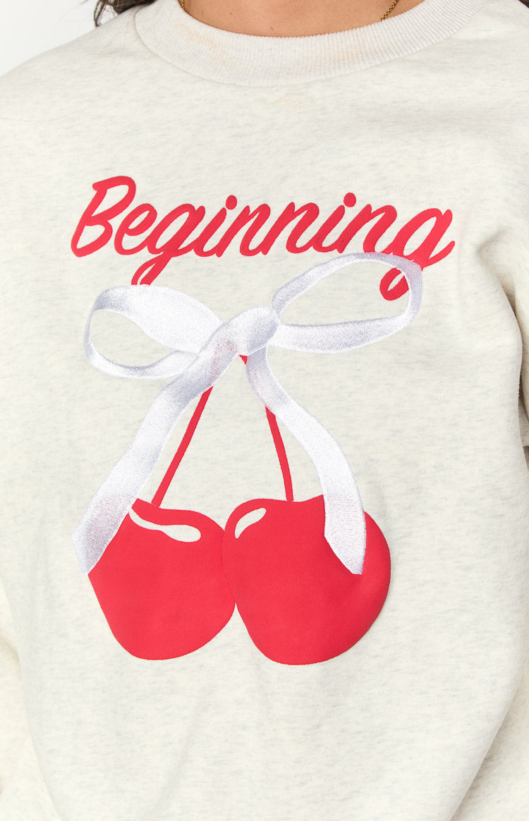 Close-up of the oat marle sweater showing the red Beginning text and cherry graphic with a white embroidered ribbon bow