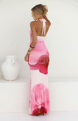 A back view of our model wearing a pale pink halter-style maxi dress with a large vibrant pink orchid print. The dress has a low-cut back with a tie at the neck perfect for your garden party.