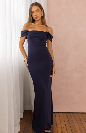 A full-length image of a model wearing a form-fitting navy blue off-the-shoulder maxi dress with a straight neckline.