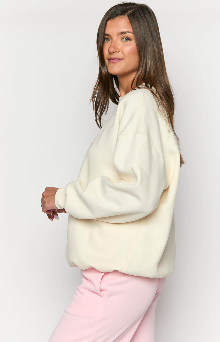 Side view of a cream crew neck sweater with a relaxed, oversized fit, dropped shoulders, and ribbed cuffs and hem