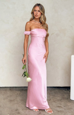 Our model is wearing a floor length maxi dress in a pink satin fabric. The dress features an off the shoulder neckline with a draped band across the chest and a subtle frill or ruffle detail on the upper edge of the sleeve. The smooth shimmering skirt is fitted and falls in a straight fluid column.