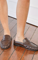 Close-up of brown leather-look slip-on loafers showing the backless mule design and penny strap detail