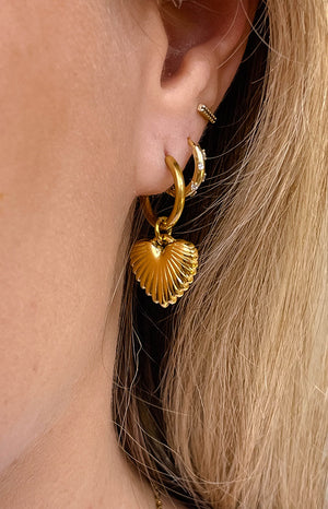 Our model earlobe showing a gold hoop earring with a gold heart-shaped charm that has a ridged shell texture. Perfect to wear in any occasion.