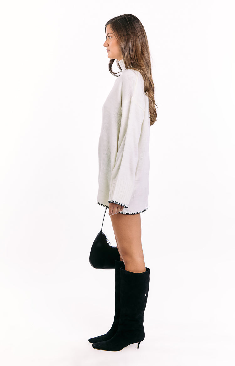 Side view of a cream oversized sweater dress with a high neck and black contrast stitching on the hem