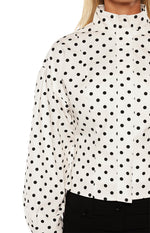  A close-up of a white and black polka dot top featuring a mock neck collar a hidden placket and a puffed long sleeve.