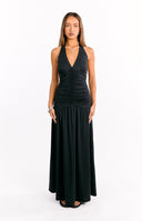 A female model wearing a black halter maxi dress with a plunging V-neckline and a ruched bodice. The skirt is loose and flowing and the model is also wearing black heeled sandals.