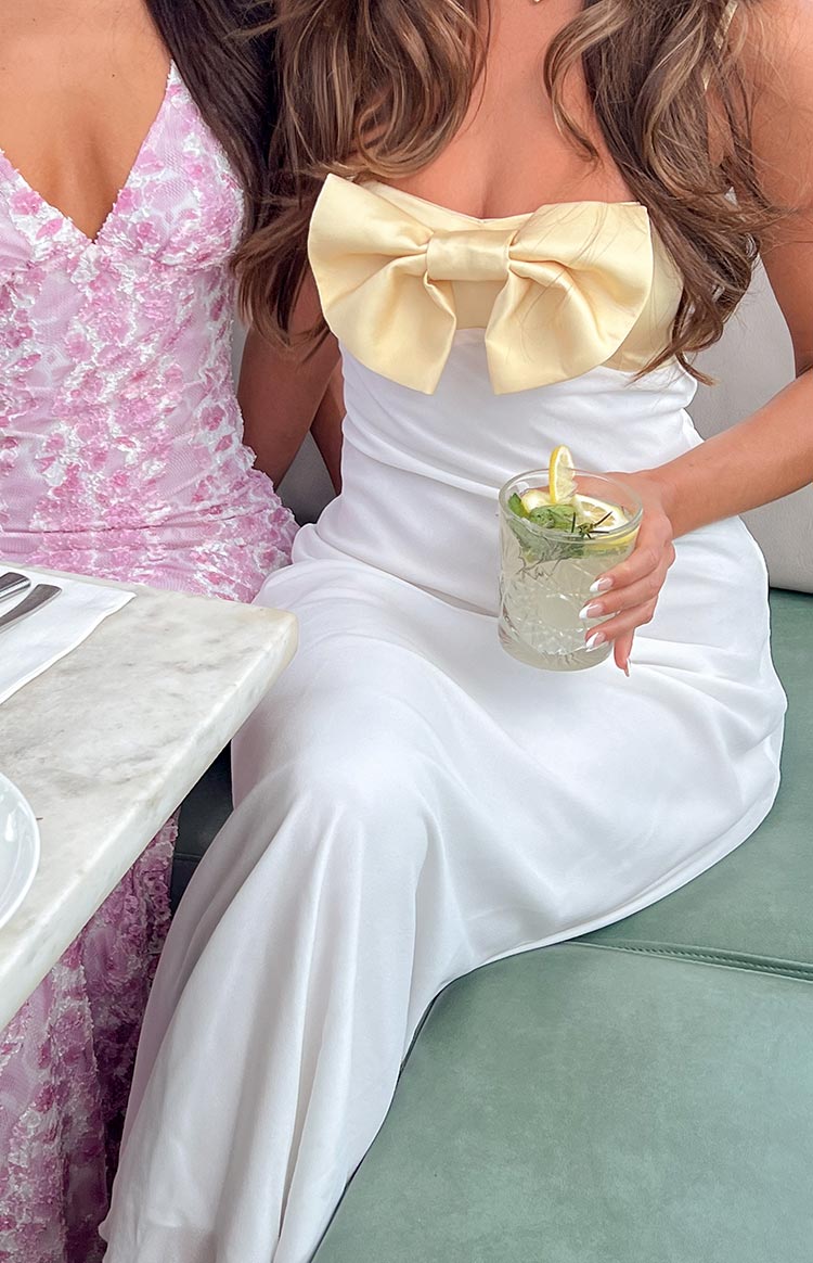 Yellow satin bow detail on the bust of a white strapless maxi dress with a flowing skirt