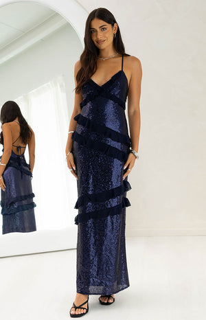 Our model wearing a floor-length maxi dress in a navy blue maxi dress. This fitted gown is completely covered in shimmering sequins and is detailed with horizontal cascading sheer fabric ruffles across the bodice and skirt. It features thin spaghetti straps and a V-neckline also trimmed with ruffles.