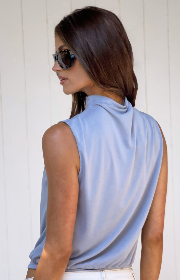 Side view of a dusty blue sleeveless top with a draped high neckline and gathered hem