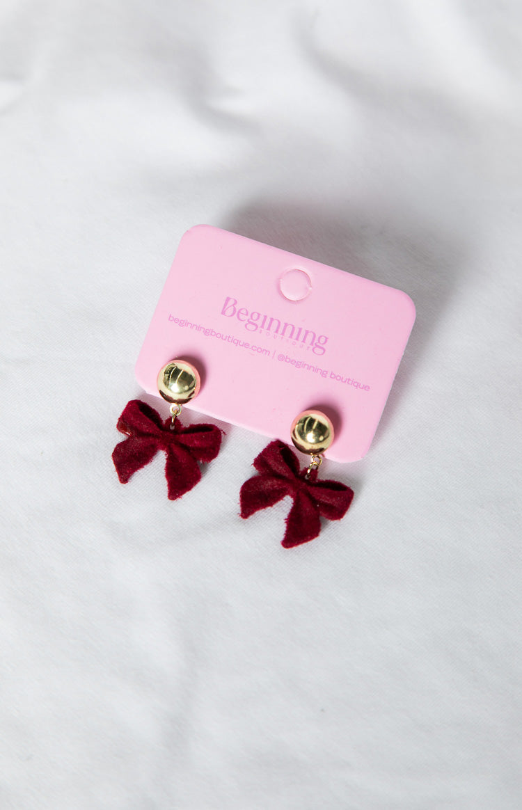 Two red velvet bow earrings with gold stud tops, presented on a pink product card