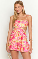 Brunette girl is wearing a mini dress with a cut out design and bright floral print. The dress has white trim details and girl is holding a white shoulder bag.