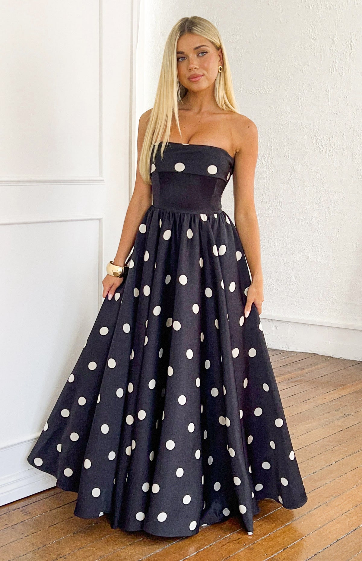 Full front view of a black polka dot satin strapless maxi dress with a fitted bodice and a full, flowing skirt
