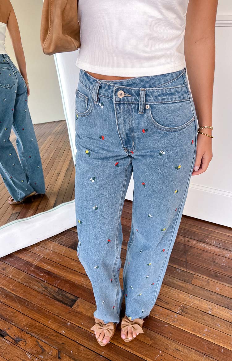 Close-up of light blue embroidered jeans with colourful strawberry and flower patterns, showing the straight leg and distressed hem