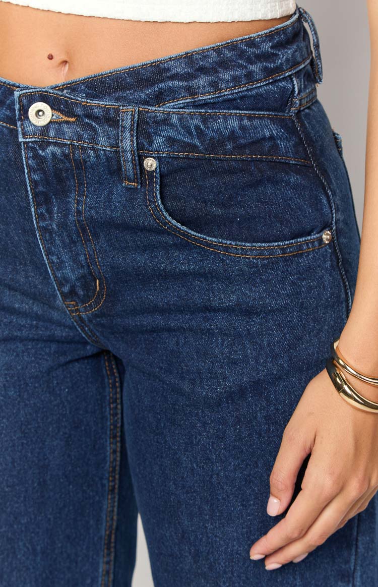 Close-up of the dark blue denim jeans featuring the uneven crossover waistline, single button closure, and orange contrast stitching