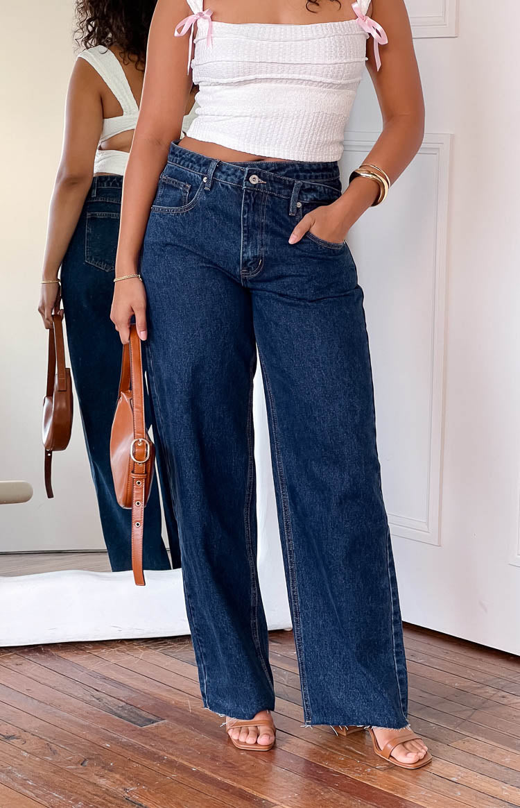 Front view of dark blue wide-leg jeans with an uneven crossover waist and raw hem