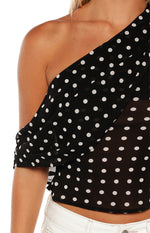 A close-up of a sheer black one-shoulder top with a white polka dot pattern highlighting the draped short sleeve.