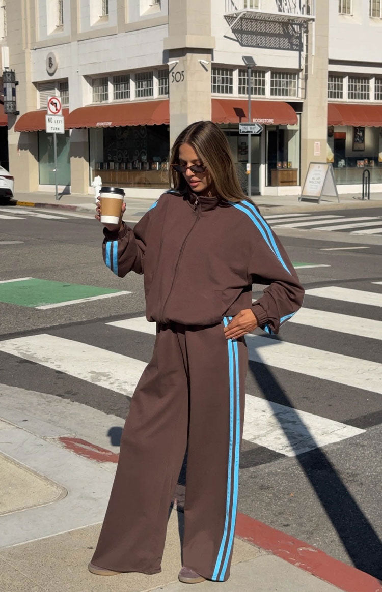 Chocolate brown wide-leg track pants with double blue side stripes, worn with a matching high-neck zip-front jacket