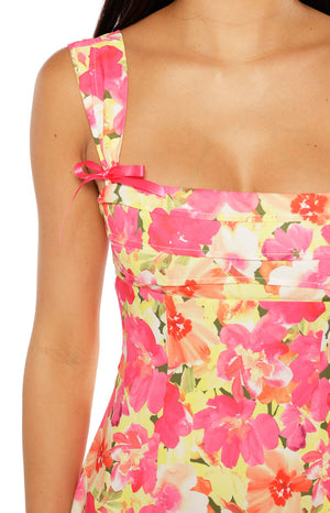A close-up of a yellow pink and orange floral print mini-dress highlighting the square neckline and the shoulder strap tied with a thin pink ribbon.