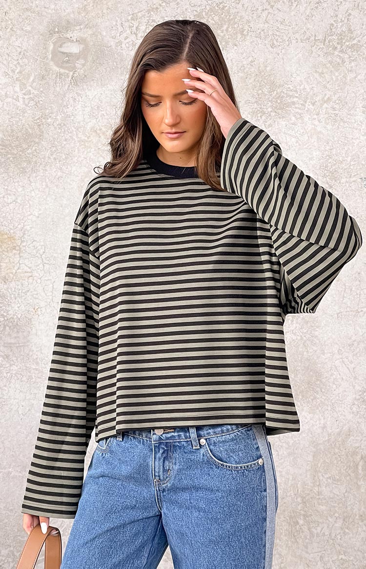 Black and light olive green horizontal striped long-sleeve top with a black crew neck, paired with blue straight-leg jeans. A brown shoulder bag is held.