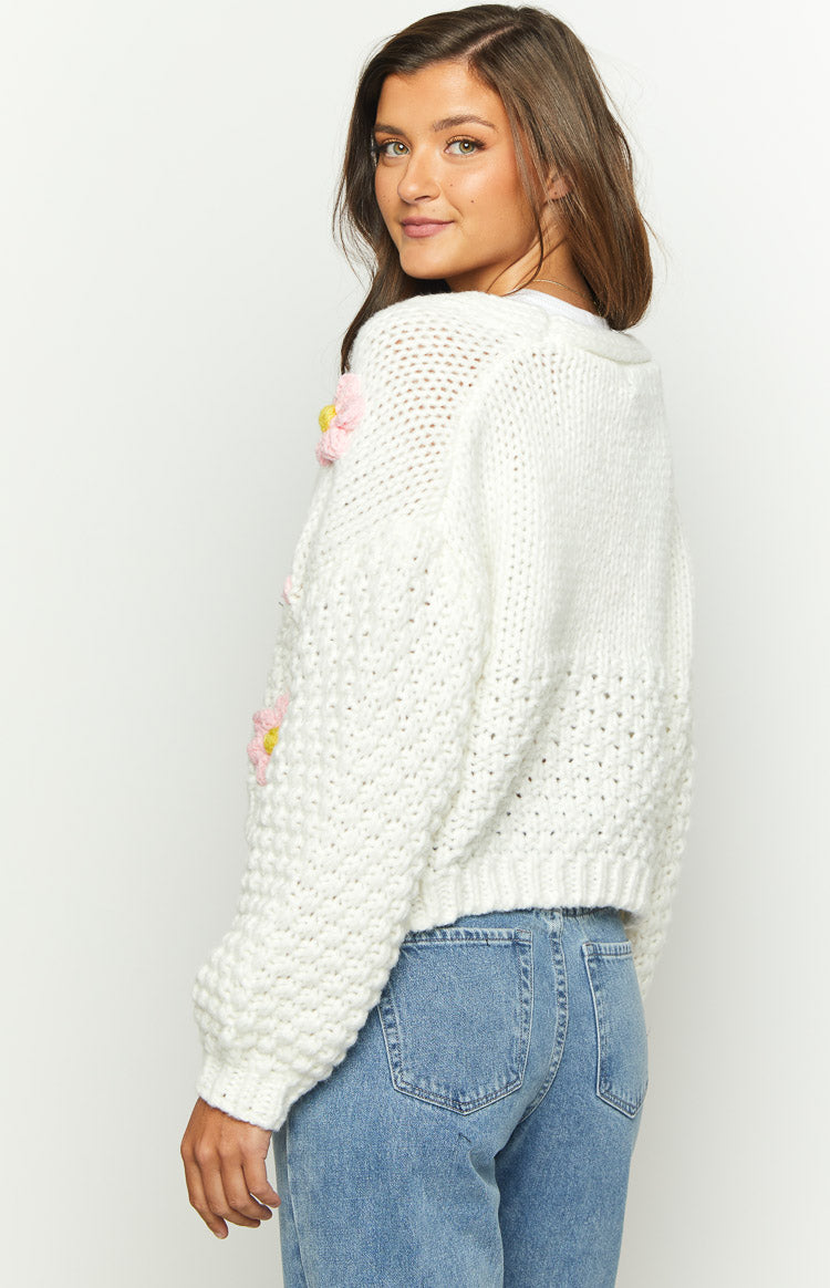 Side view of a white knit cardigan with textured stitches, cropped length, and pink and yellow flower appliques on the sleeve