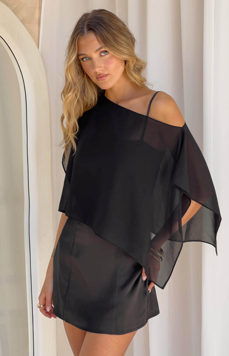 Black satin mini dress with a sheer one-shoulder cape and a thin spaghetti strap on the other shoulder