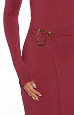 A close-up view of a long-sleeve maxi dress in a burgundy color. The smooth fitted fabric of the dress is gathered at the waist by a large asymmetric gold-toned metal buckle accent. The skirt features a diagonal wrap slit overlap. This maxi visible over the skirt wearing long sleeves that extend past the wrist.