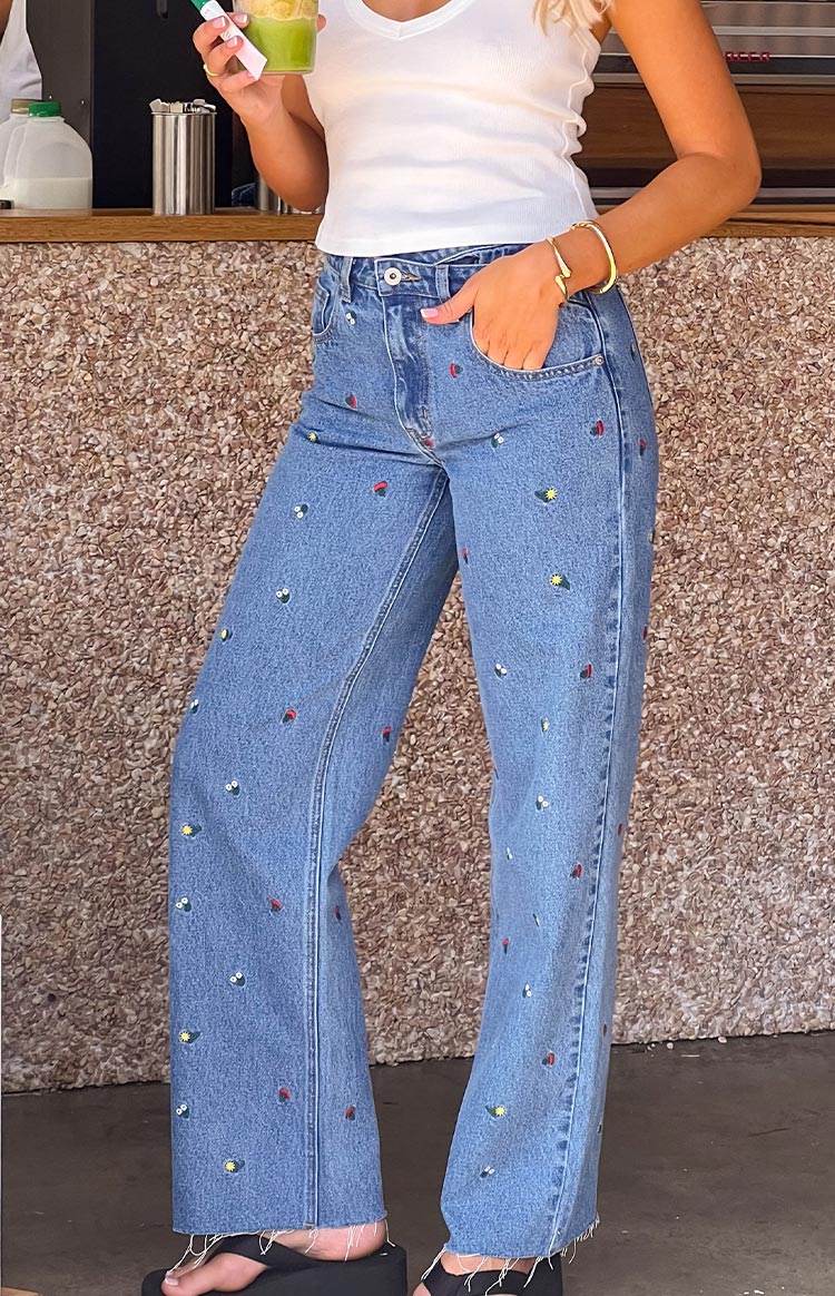 Full length view of blue denim straight-leg jeans featuring small colourful embroidered motifs and a raw hem