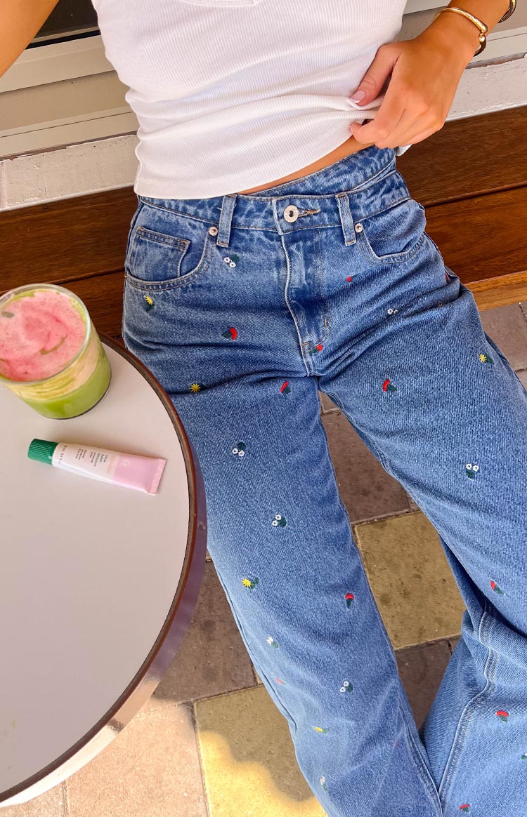 Close-up of the blue denim jeans showing the front pockets and scattered colourful embroidered fruit and flower motifs